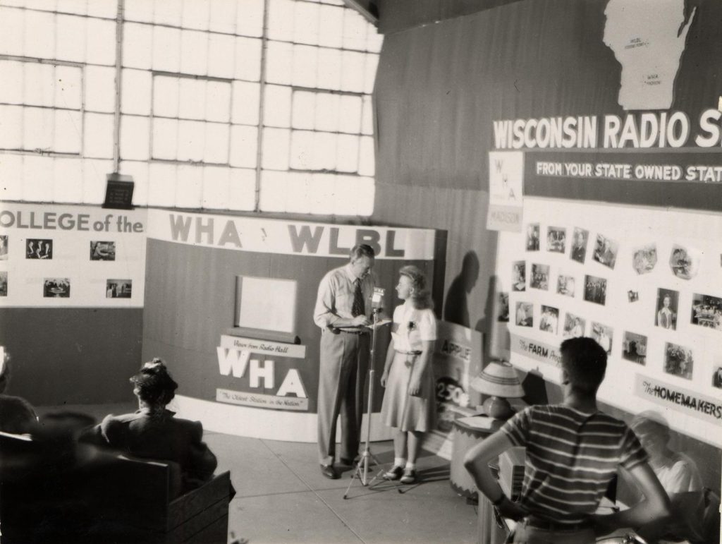 A man and woman stand at microphone in the midst of a WHA display at a state fair. Archives